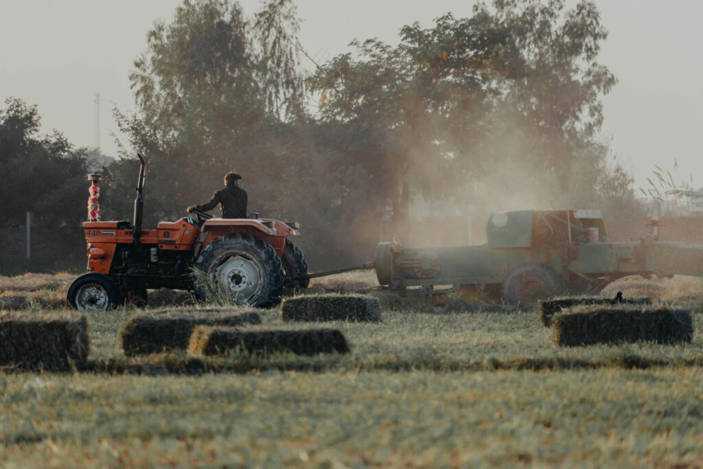 A farmer driving a tractor through a dusty rural field, harvesting hay bales.