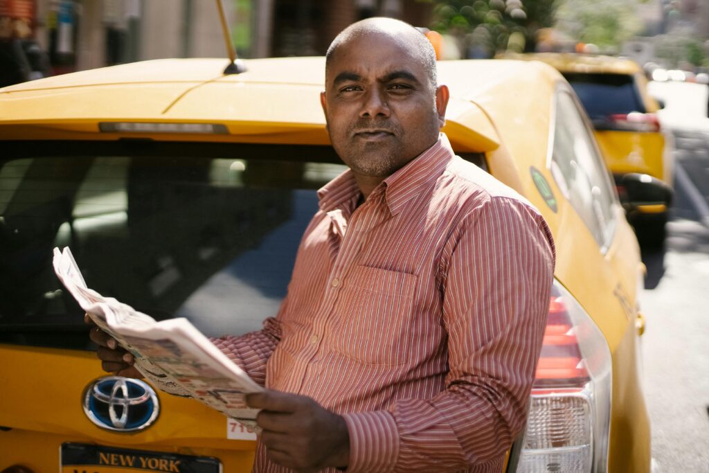 A taxi driver reads a newspaper next to his cab on a sunny urban street.
